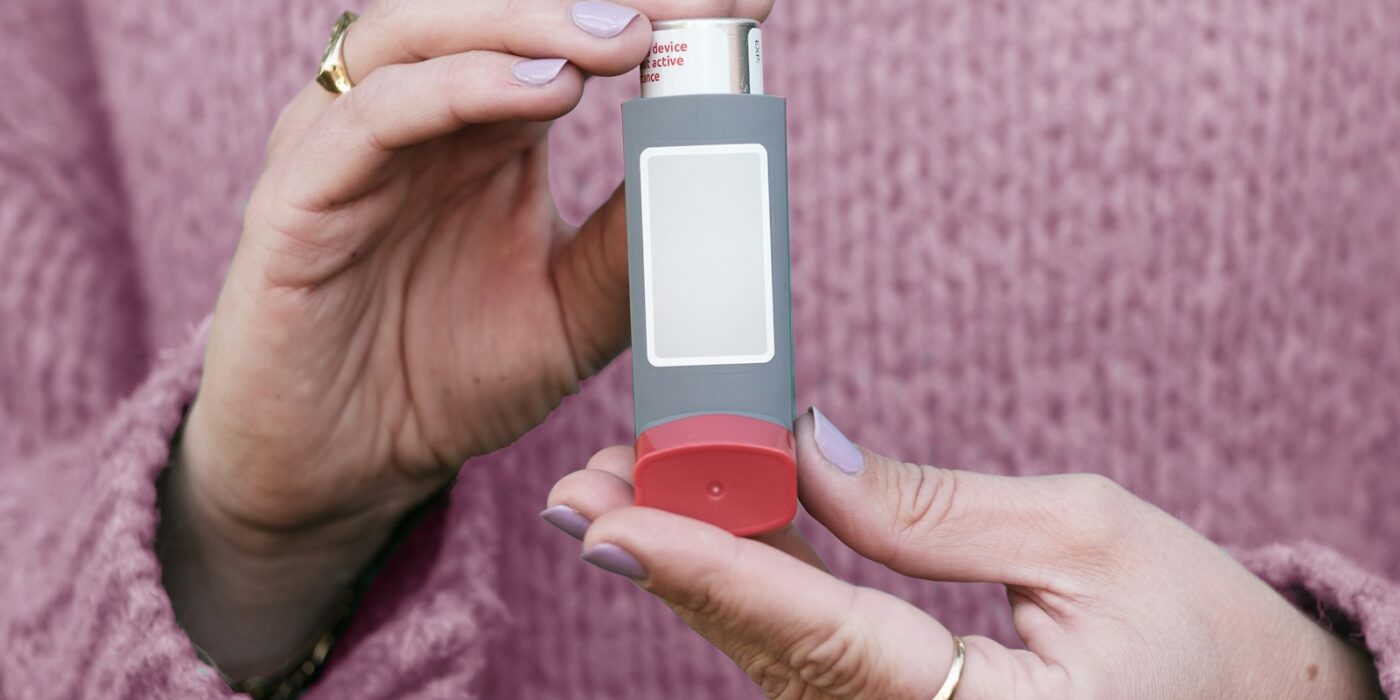 Two hands holding a gray metered-dose inhaler with a blank label in front of a pink knit sweater