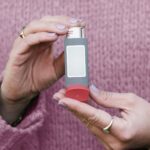 Two hands holding a gray metered-dose inhaler with a blank label in front of a pink knit sweater