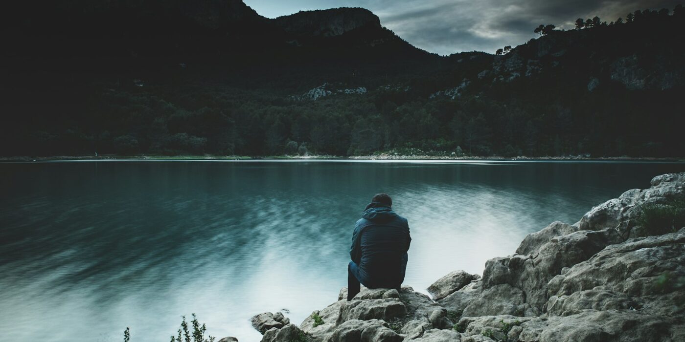A man sitting alone on lakeside rocks at dusk, seen from behind, looking across calm water toward dark hills