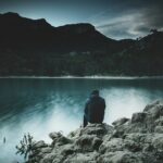 A man sitting alone on lakeside rocks at dusk, seen from behind, looking across calm water toward dark hills