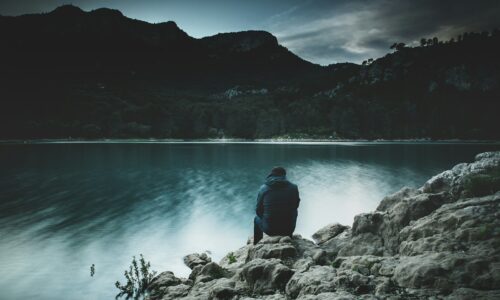 A man sitting alone on lakeside rocks at dusk, seen from behind, looking across calm water toward dark hills