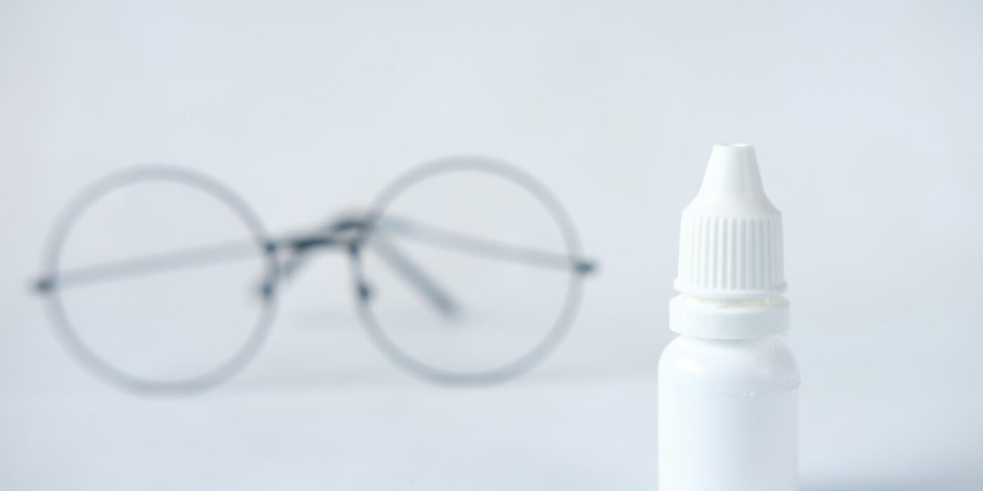 A white unlabeled eye drop bottle on a light surface with a pair of round glasses blurred in the background