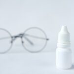 A white unlabeled eye drop bottle on a light surface with a pair of round glasses blurred in the background