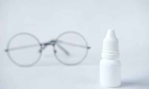 A white unlabeled eye drop bottle on a light surface with a pair of round glasses blurred in the background