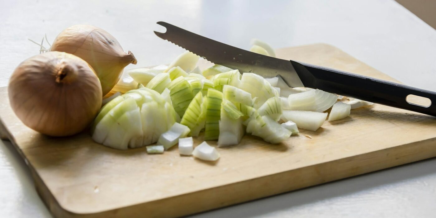 Whole and chopped yellow onions with a serrated knife on a wooden cutting board on a white table