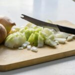 Whole and chopped yellow onions with a serrated knife on a wooden cutting board on a white table