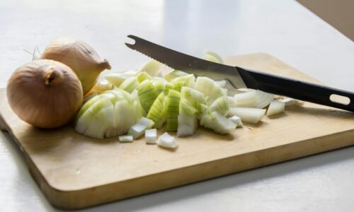 Whole and chopped yellow onions with a serrated knife on a wooden cutting board on a white table
