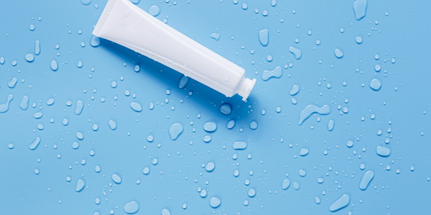 A blank white cream tube lying on a light blue surface scattered with water droplets