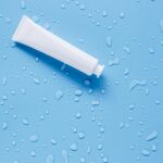 A blank white cream tube lying on a light blue surface scattered with water droplets